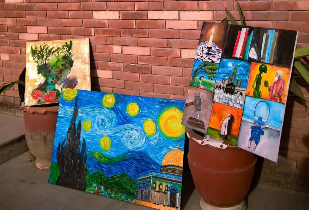 Three paintings by Hafsa Imran leaning against a brick wall: the gold-leaf lotus painting, the Masjid al-Aqsa night-sky painting and the nine-panel painting