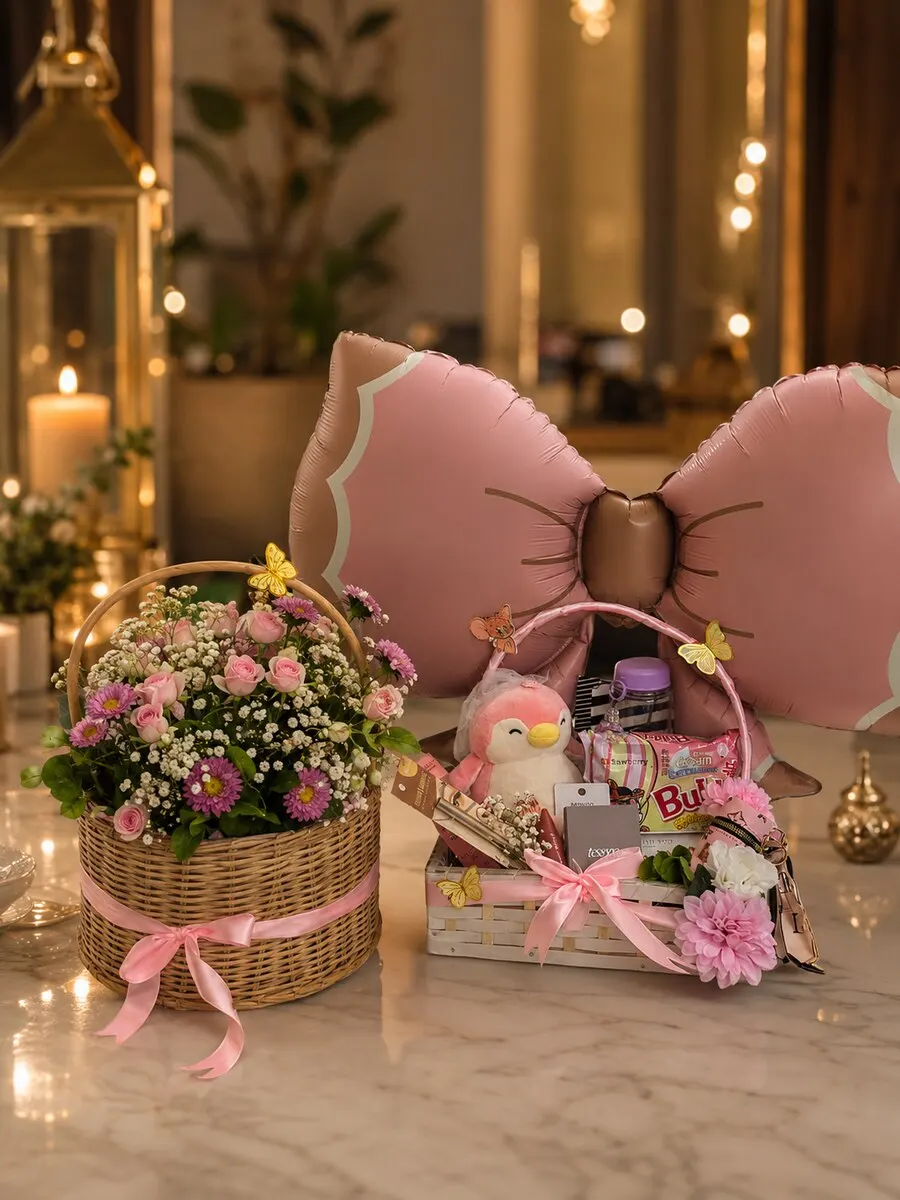 A wicker basket of fresh pink flowers beside a padded pink bow cushion and a tray of newborn baby items — handcrafted by Bloom & Beyond in Lahore