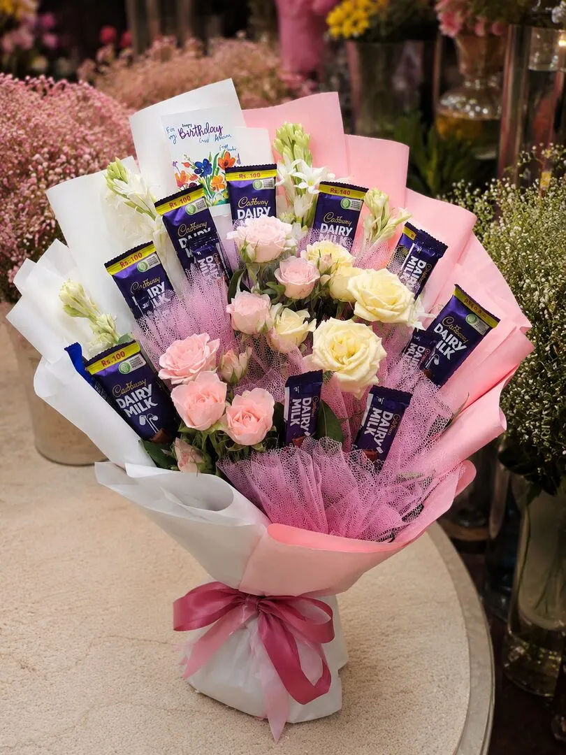 Cadbury Dairy Milk bars are arranged among soft pink and cream roses with a 'Happy Birthday' card — handcrafted by Bloom & Beyond in Lahore