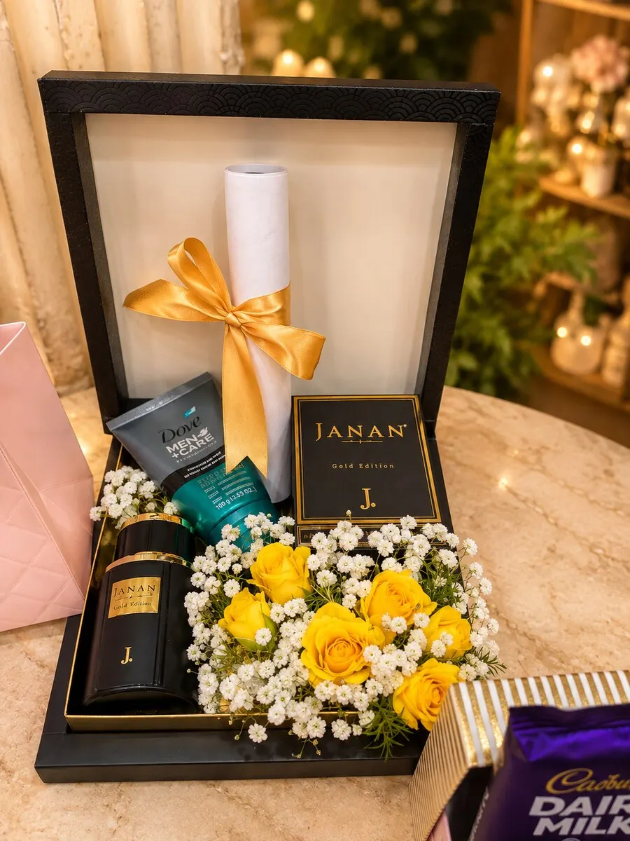 A black presentation gift box holding JANAN men's grooming products, a scroll tied with gold ribbon and fresh yellow roses — handcrafted by Bloom & Beyond in Lahore