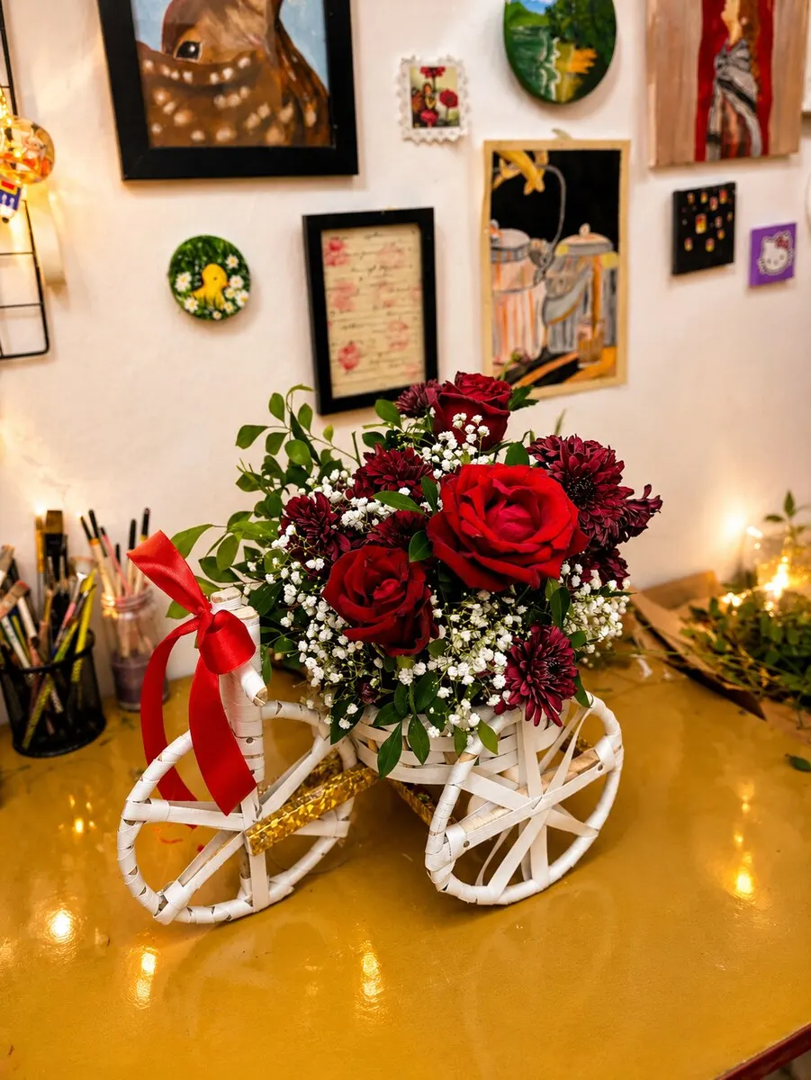 Deep red roses and burgundy blooms with baby's breath arranged in a white bicycle cart tied with red ribbon — handcrafted by Bloom & Beyond in Lahore