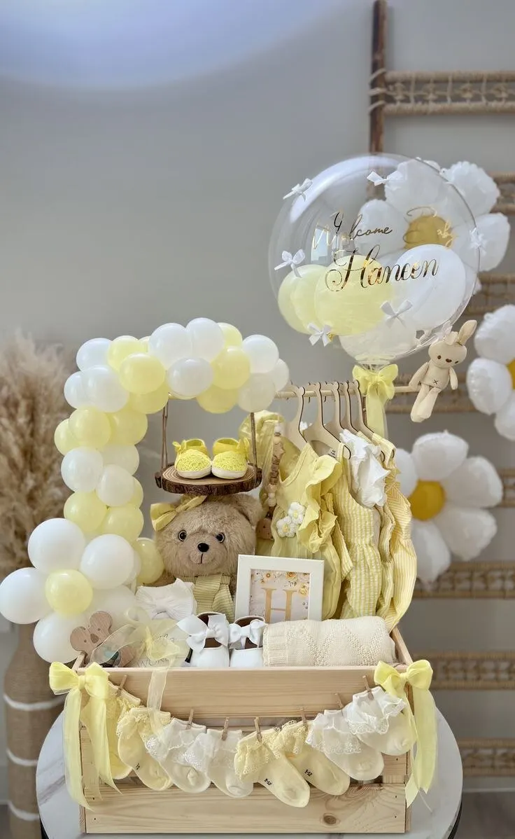 A cream and yellow balloon arch over a wooden crate of newborn clothes on small hangers with a teddy bear and a personalised welcome balloon — handcrafted by Bloom & Beyond in Lahore