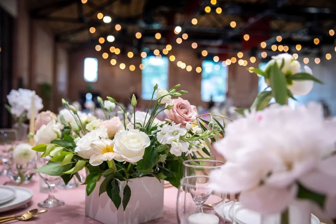White roses and peonies in a white block vase on a laid dinner table, string lights behind