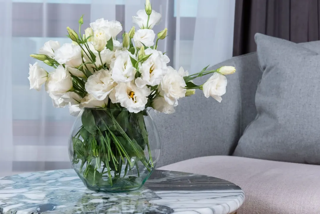 White lisianthus in a round glass vase on a marble side table beside a grey sofa