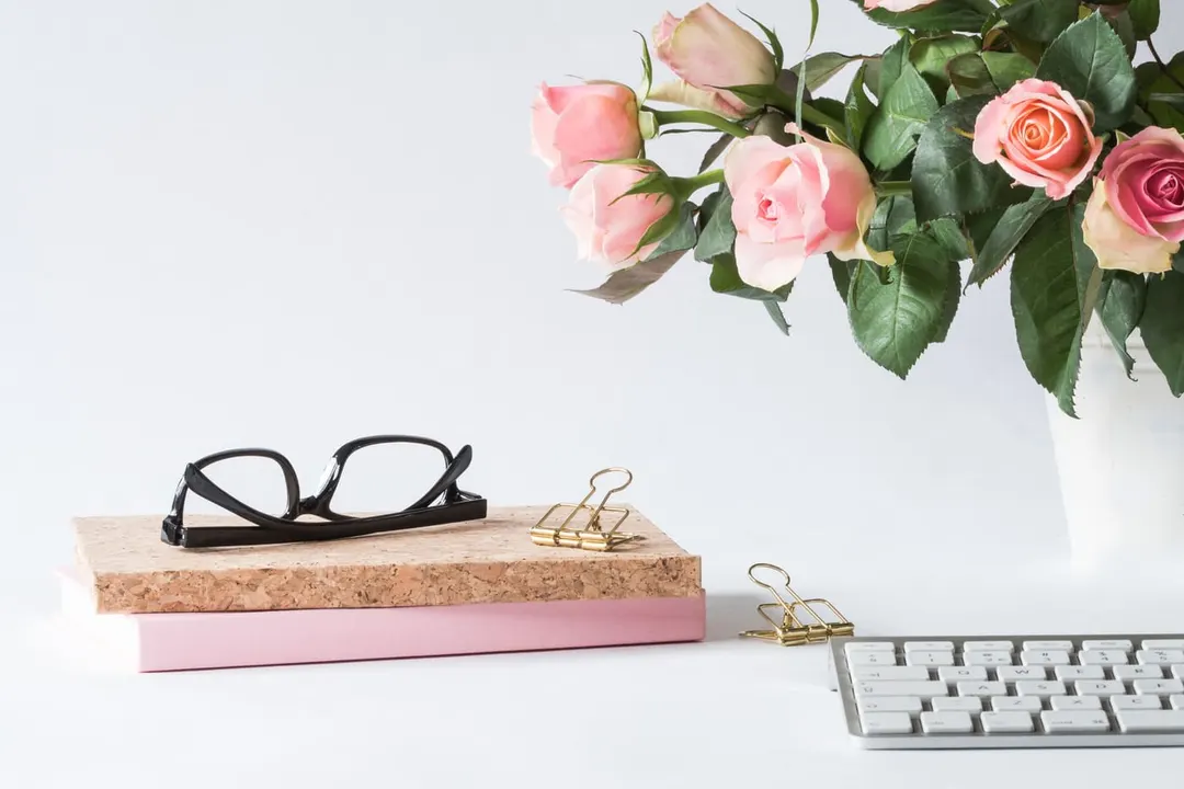 Pink roses in a white vase on a desk beside a keyboard, glasses and notebooks