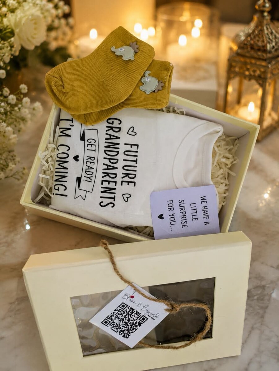 A cream gift box holding a white bodysuit printed Future Grandparents Get Ready I'm Coming, with mustard knitted socks with grey elephants and a lilac announcement card — handcrafted by Bloom & Beyond in Lahore