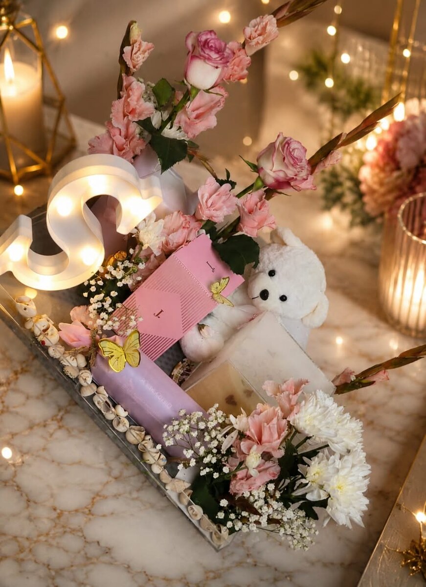 A marble tray with a lit marquee initial letter, pink roses and white chrysanthemums, a white teddy bear and a wrapped gift box — handcrafted by Bloom & Beyond in Lahore