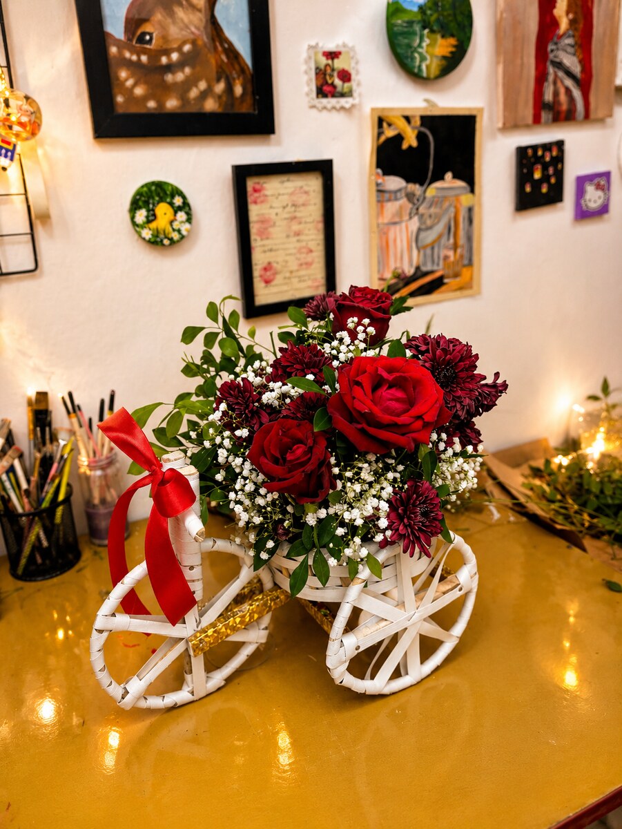 Deep red roses and burgundy blooms with baby's breath arranged in a white bicycle cart tied with red ribbon — handcrafted by Bloom & Beyond in Lahore