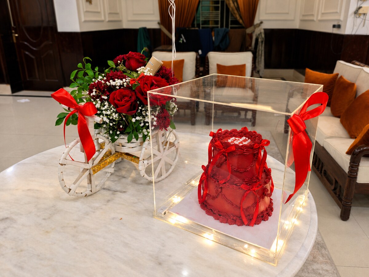 Red roses and baby's breath in a white cart beside a red heart-shaped cake under a lit acrylic dome — handcrafted by Bloom & Beyond in Lahore